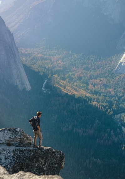 Man standing on the cliff of the gray rocks, overlooking the forest during the day
